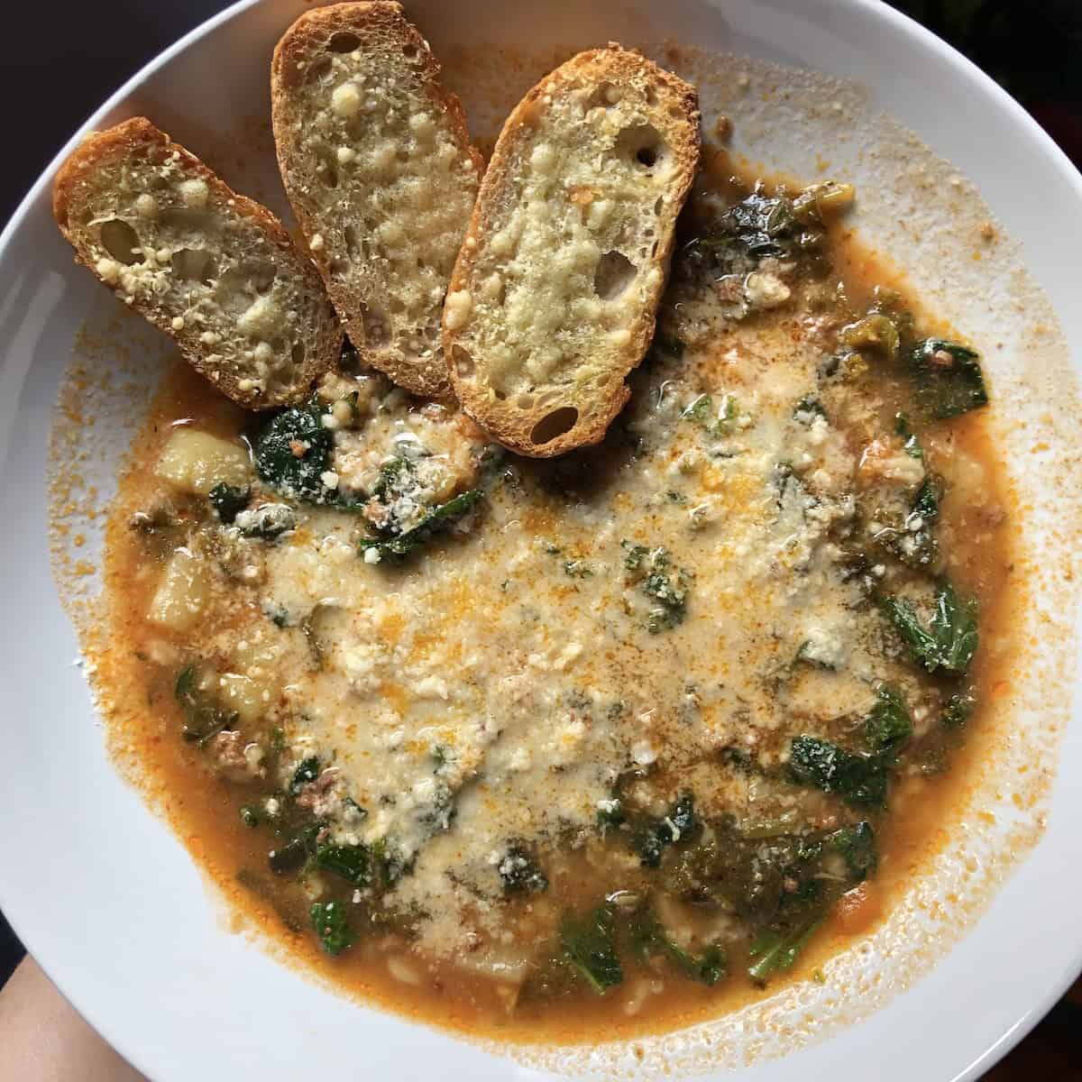 A piping hot bowl of Tuscan kale and cannelini bean soup with grated parmesan cheese and 3 garlic-parmesan bread crostini tucked into the bowl.