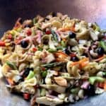 A wok filled with Mediterranean pasta salad with cooked vegetables, raw veggies, cured olives, shrimp, ham, and mozzarella cheese.