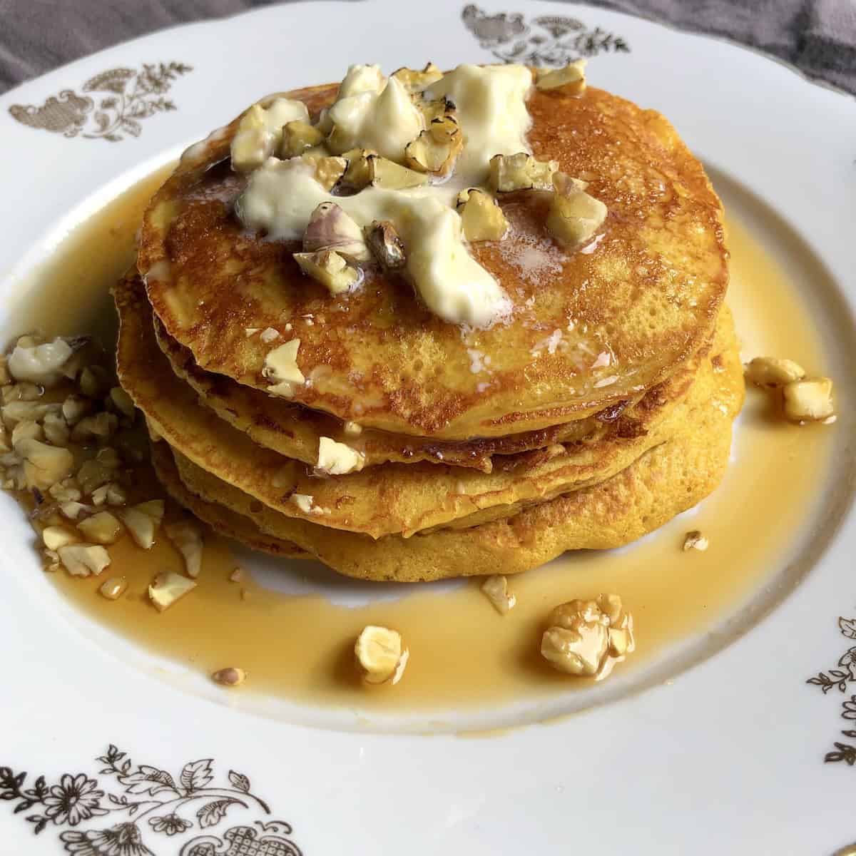 Stack of perfect fluffy pumpkin pancakes seasoned with homemade pumpkin spice with butter melting on top, pure maple syrup dripping off the stack and surrounding them on the plate with toasted chestnuts sprinkled all over. 
