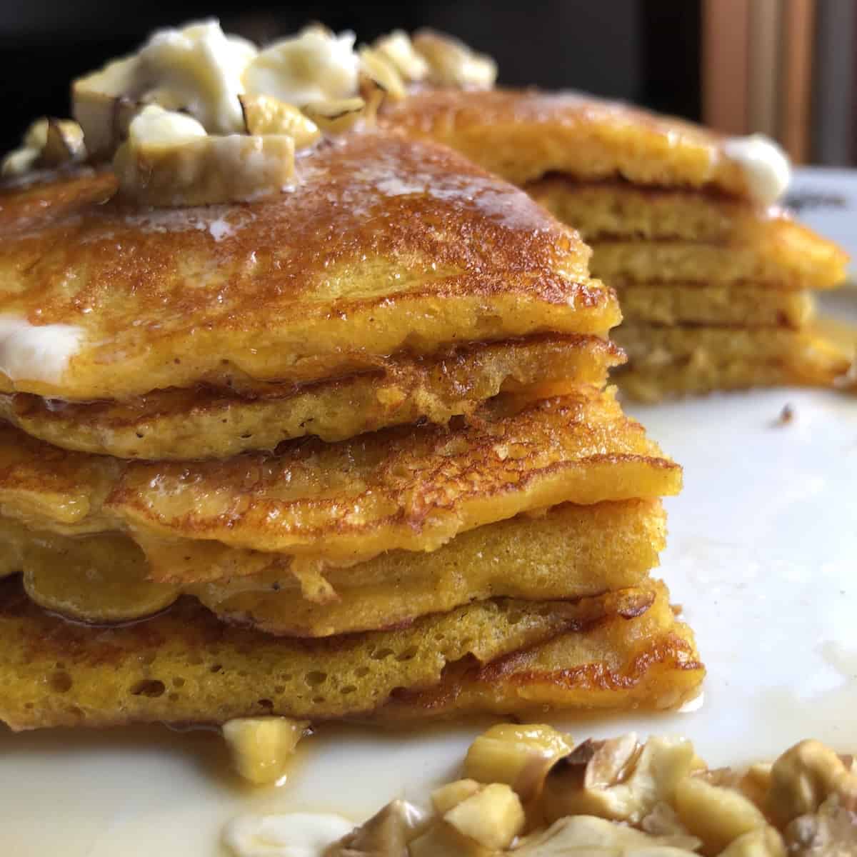 Side view of stacked fluffy pumpkin pancakes with maple syrup, melted butter and toasted chestnuts showing the best fluffy pumpkin pancakes recipe for light and fluffy pumpkin pancakes from scratch.
