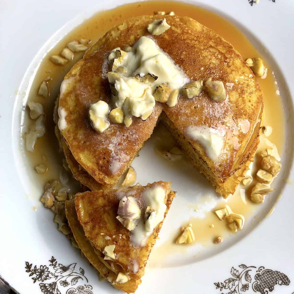 Top down view of a stack of fluffy pumpkin pancakes with maple syrup, butter and toasted chestnuts with wedge removed showing light and fluffy interior texture for the best pumpkin pancakes recipe from scratch.