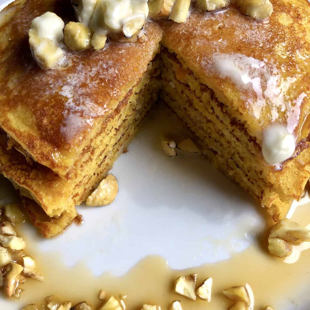 Stack of fluffy pumpkin pancakes with maple syrup, butter and toasted chestnuts with wedge removed showing light and fluffy interior texture for the best pumpkin pancakes recipe from scratch.