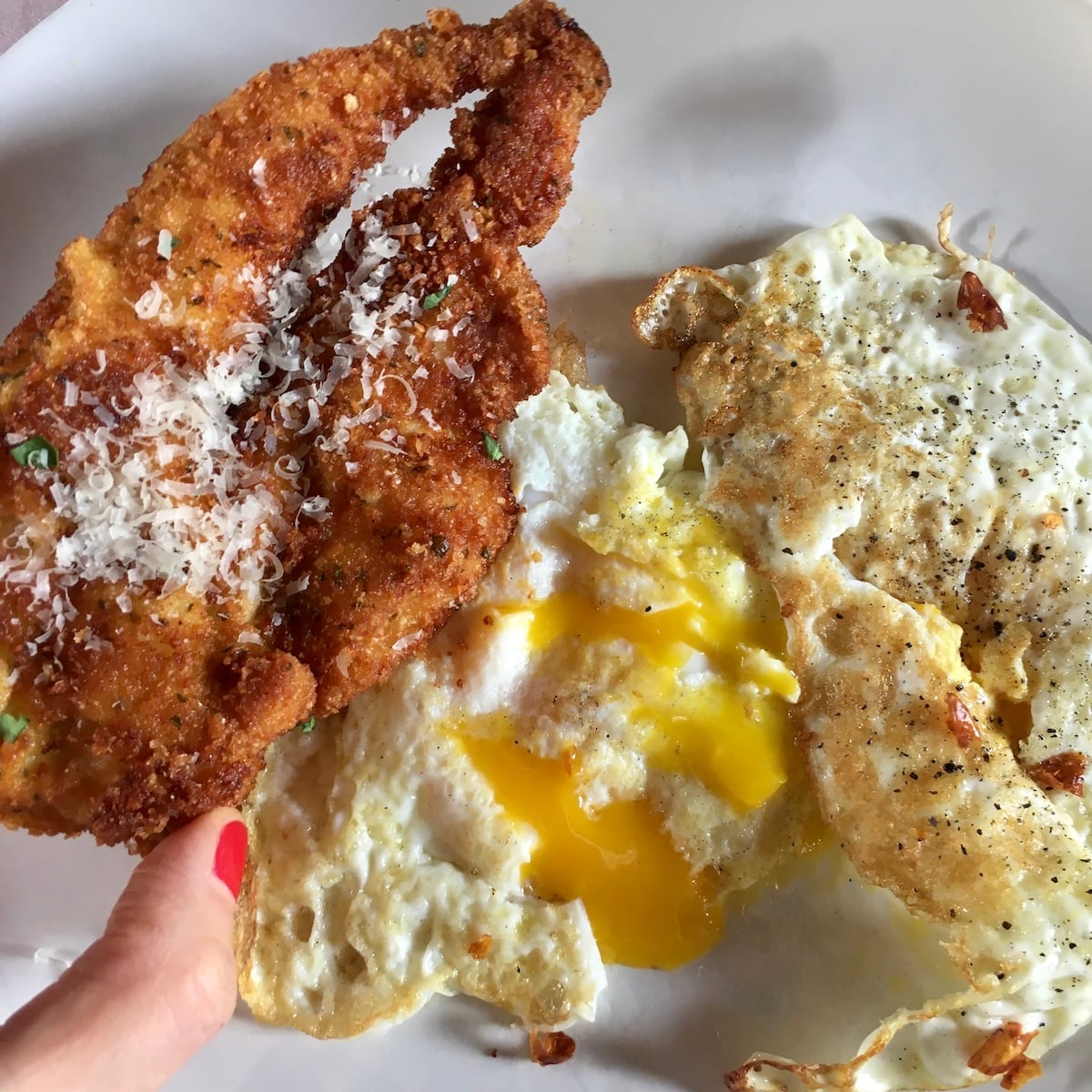 My hand holding a perfectly golden brown crispy basil-parmesan panko-crusted chicken breast over a plate with eggs over medium with yellow yolks and cooked whites.