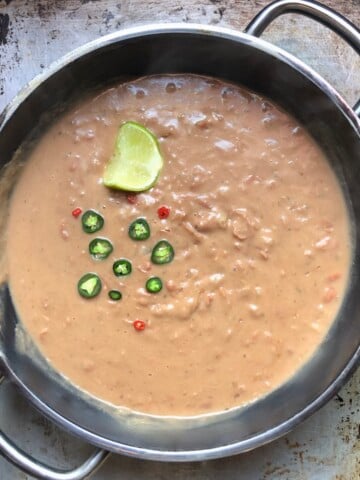 A saute pan with the creamiest refried beans garnished with a a few sliced fresh jalapeños, chili pequin, and a lime wedge.