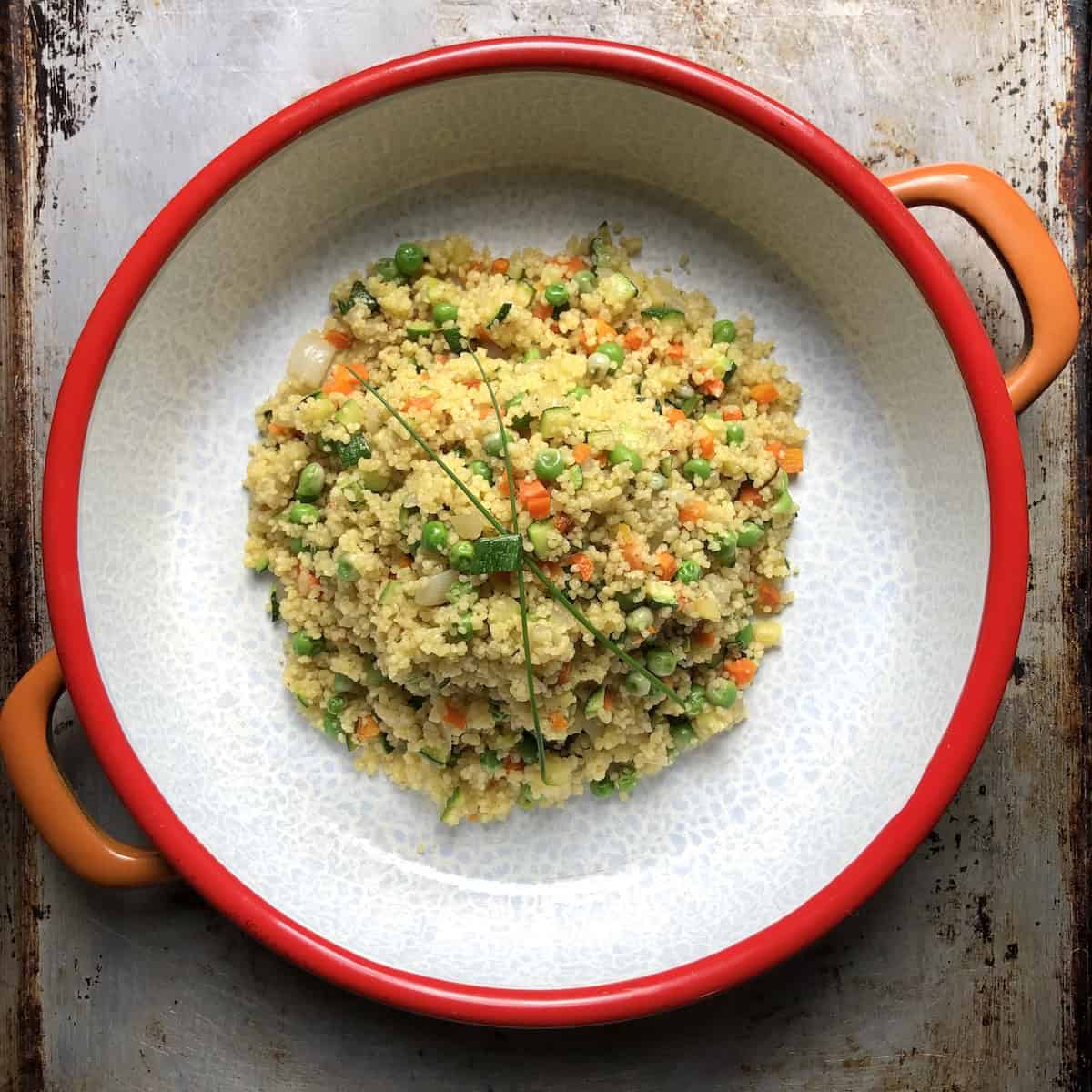 Authentic homemade Italian Mediterranean vegetable couscous served in vintage round dish featuring perfectly cooked couscous with sweet green peas, caramelized diced carrots, zucchini, and onions, garnished with fresh chive sprigs and traditional Italian seasonings.