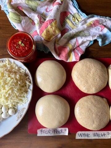 Side-by-side comparison of six pizza dough balls (two each of bread flour, whole wheat flour, and 00 flour) with a serving platter of freshly grated whole milk mozzarella, mini mozzarella balls, and grated grana padano cheese, alongside a mason jar of homemade pizza sauce.