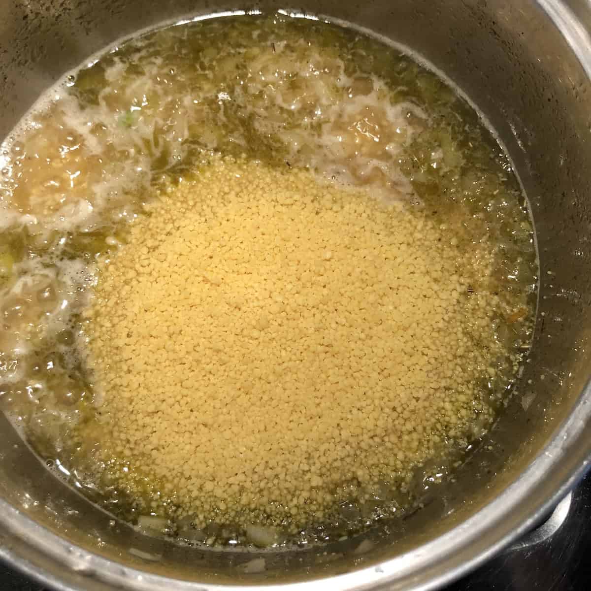 Adding dry couscous to boiling chicken stock with sautéed onions, butter and Mediterranean herbs in a pot, demonstrating the proper cooking technique for authentic Italian vegetable couscous that ensures perfect texture and flavor absorption.