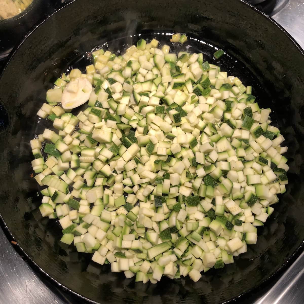 Fresh diced zucchini being added to a hot cast iron skillet with smashed garlic for authentic Italian Mediterranean vegetable couscous recipe, showcasing the proper technique for cooking vegetables separately to build flavor in homemade couscous.