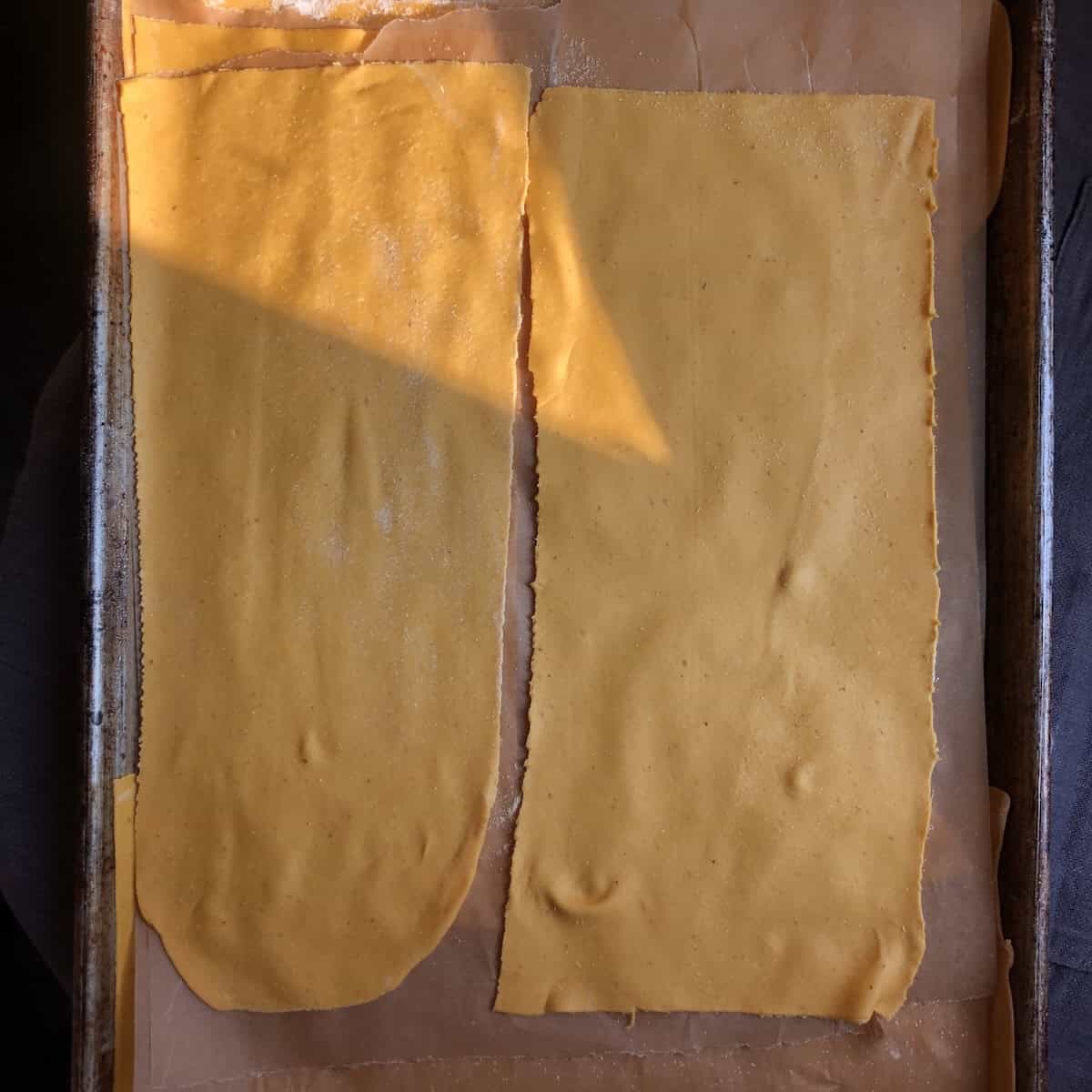 Homemade pumpkin lasagna noodles fresh pasta sheets layered with parchment paper dusted with semolina flour on baking sheet for freezing or resting before cooking.