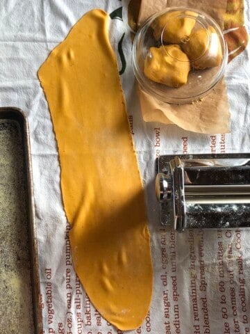 Homemade pumpkin pasta dough with lasagna sheets and Atlas Marcato 150 pasta machine next to it with 3 pumpkin pasta dough balls covered in the background.