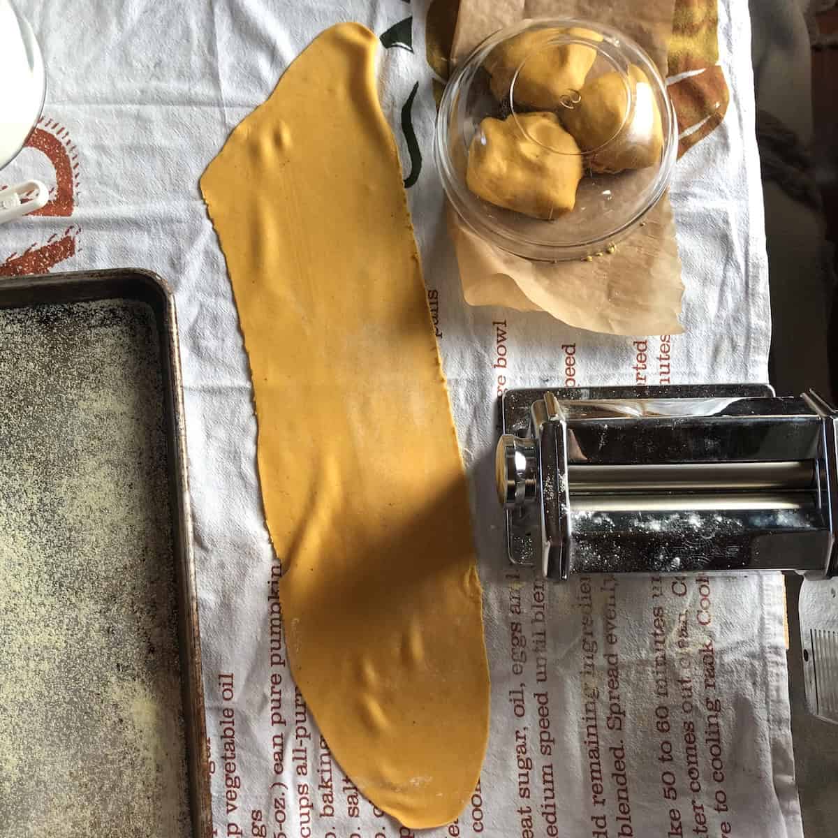 Homemade pumpkin pasta dough with lasagna sheets and pasta machine next to it with 3 pumpkin pasta dough balls covered in the background.