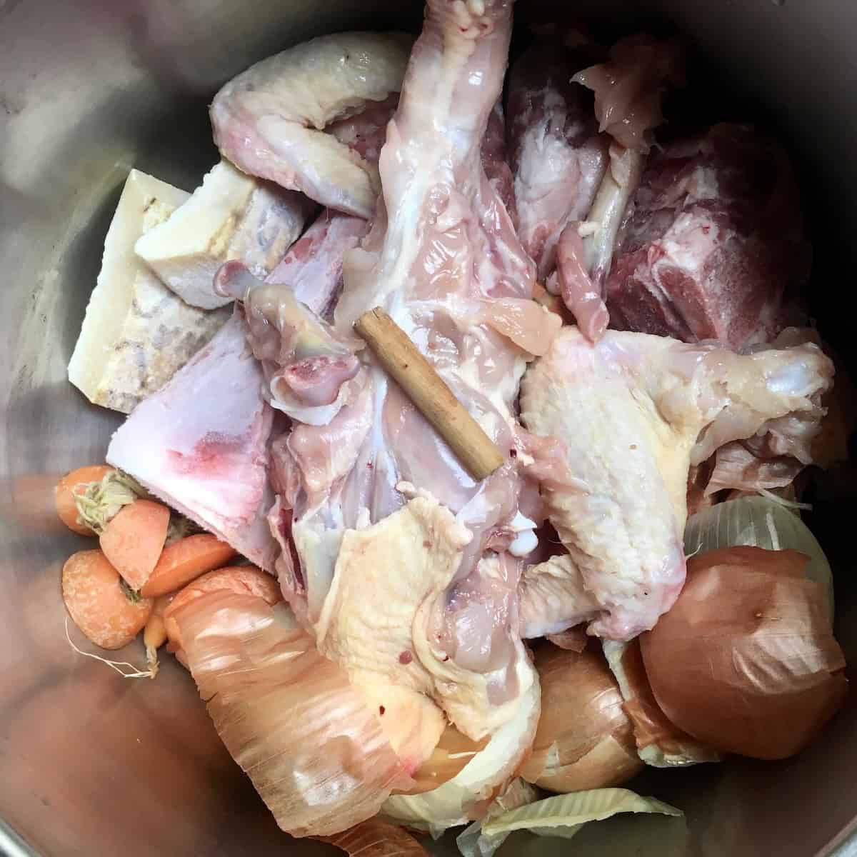 Pressure cooker filled with chicken bones, vegetables, peppercorns, salt, and grana padano cheese rind and 2 inch piece of cinnamon stick for homemade chicken broth recipe before adding water showing how to make chicken broth.