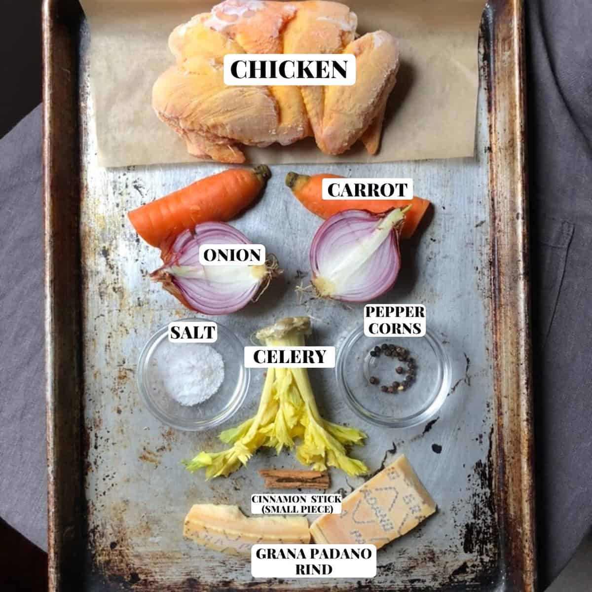 Homemade chicken broth ingredients including chicken, carrots, celery, onion, and sea salt, black peppercorns, leftover aged grana padano rind and 1-inch piece of cinnamon stick (Italian secret ingredients for the best chicken broth) arranged on a sheet pan in the shape of a face.