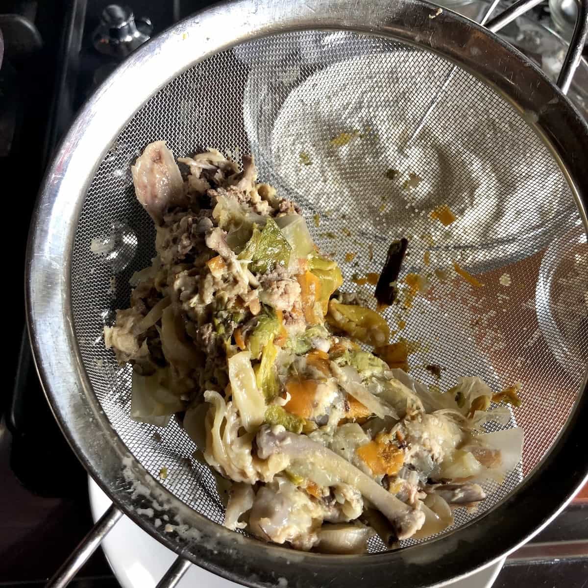Fine mesh strainer being used to strain homemade chicken broth leaving unusable cooked vegetables behind showing how to make chicken broth final step.