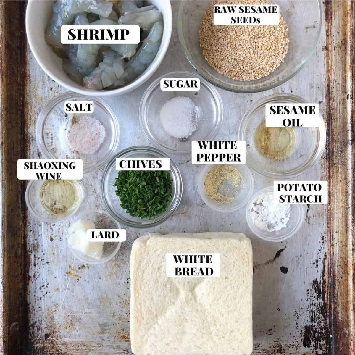 Easy shrimp toast ingredients labeled on sheet pan including raw shrimp, sesame seeds, white bread, and seasonings for homemade Chinese shrimp toast.
