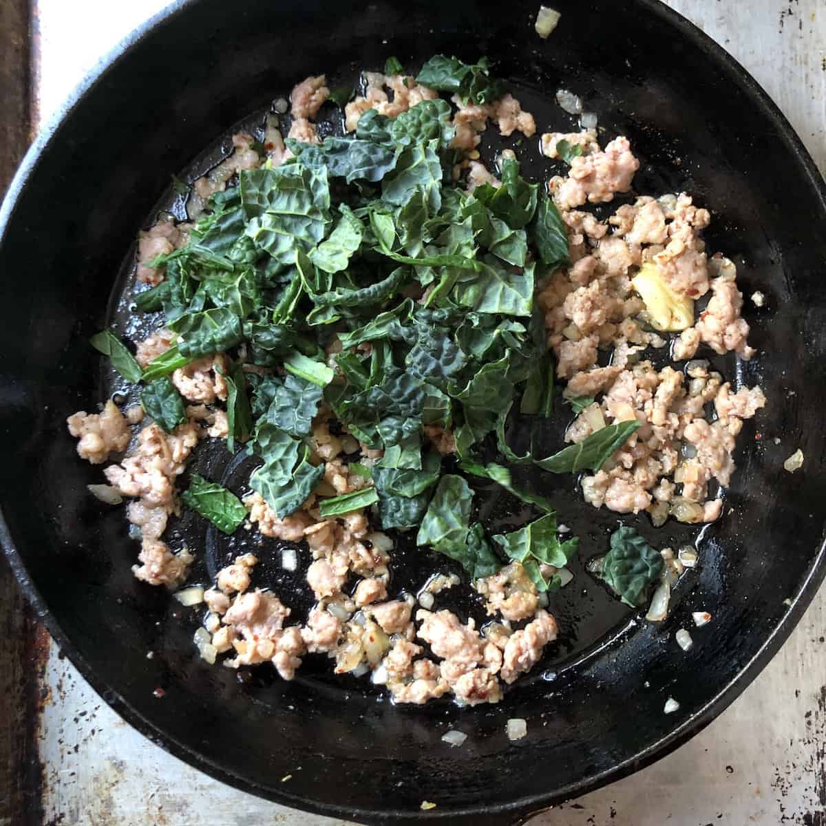 Chopped kale added to sausage mixture in skillet for sausage kale pasta, a key step in pumpkin pasta recipes.
