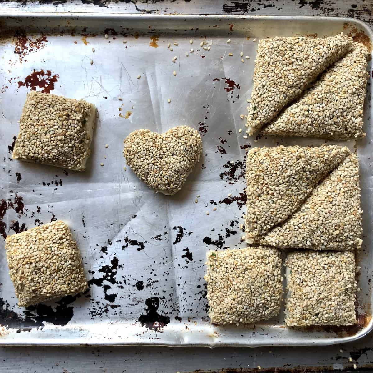 Fully assembled heart-shpaed shrimp toasts (also triangles and squares) with sesame seed coating on sheet pan ready to fry for golden crispy shrimp toast recipe.
