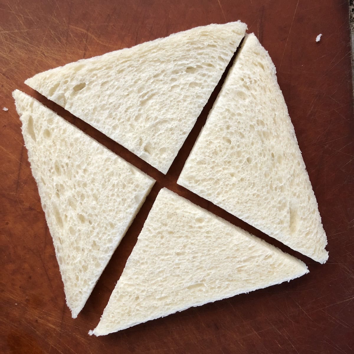 Crustless white sandwich bread cut into four triangles on cutting board for homemade shrimp toast recipe.
