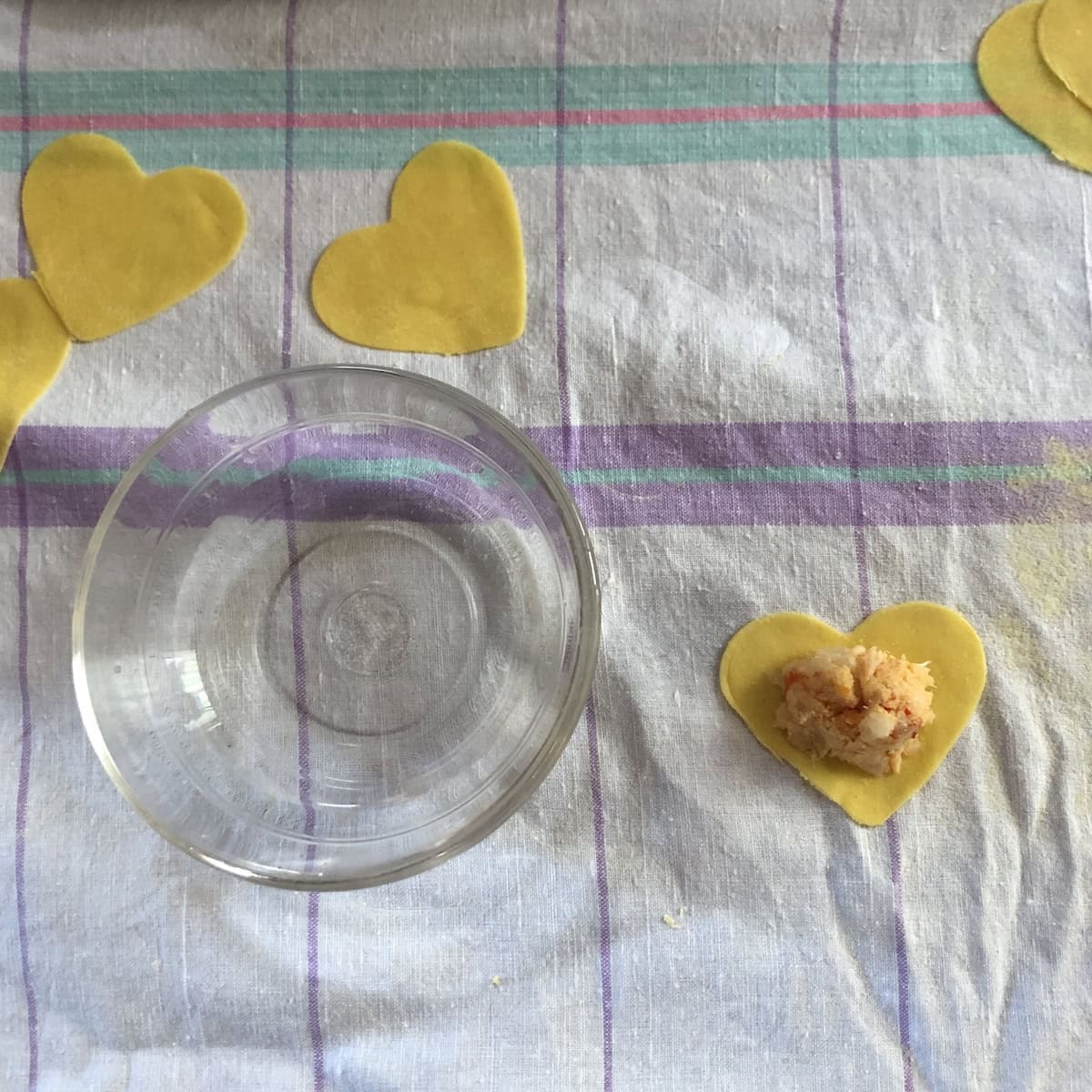 Bowl of water next to heart shaped ravioli with spoonful of lobster ravioli filling in the middle ready for top layer of pasta.
