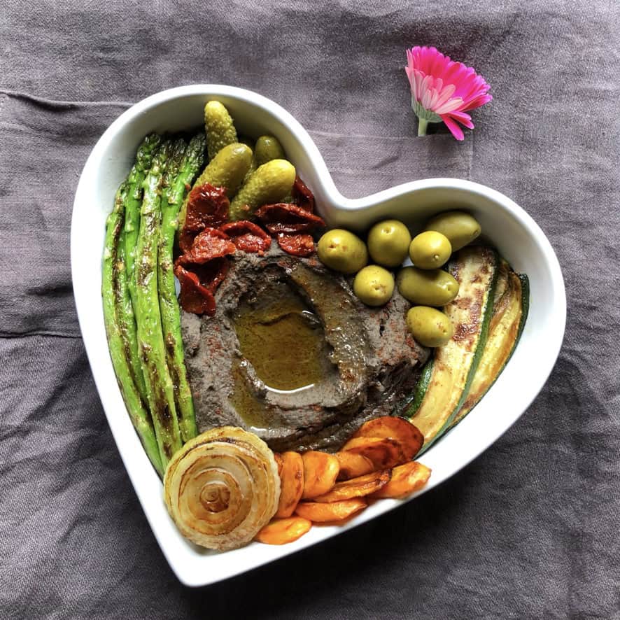 Smooth black chickpea hummus in a white heart-shaped casserole dish with a well of extra virgin olive oil, surrounded by caramelized onions, pan-seared asparagus, sun dried tomatoes, green olives, cornichon pickles, sauteed carrot medallions, and golden brown zucchini. A beautiful meatless lenten snack or meal.