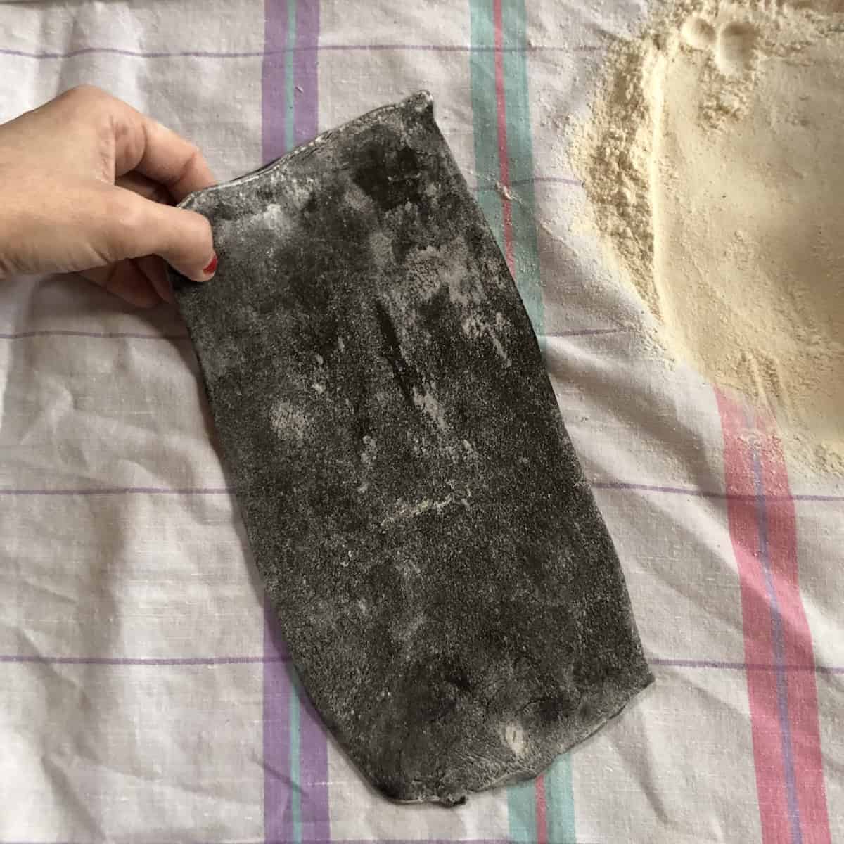 Black squid ink pasta dough rectangle after narrower setting pass, ready to roll through thinner settings for fresh homemade nero di seppia pasta.
