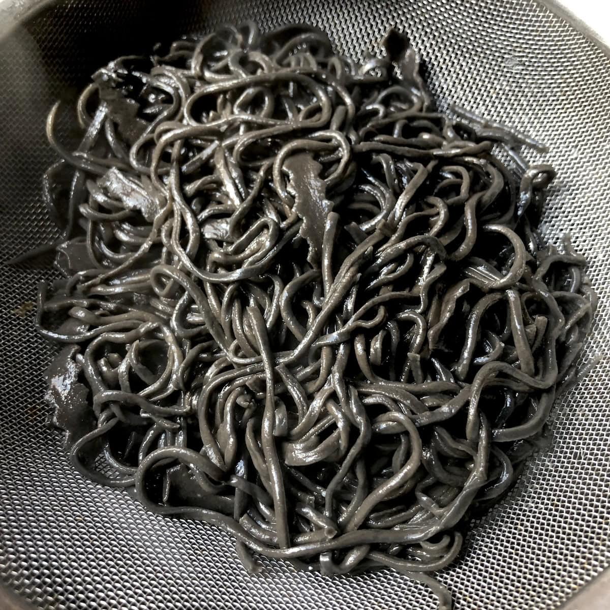 Cooked homemade black squid ink spaghetti pasta in fine mesh strainer ready for sauce, fresh nero di seppia noodles for seafood pasta.
