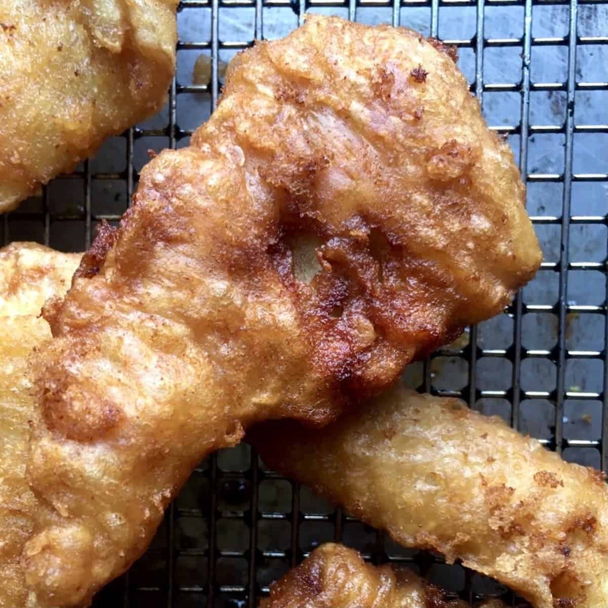 Super crispy golden brown British style fried fish filet resting on a wire rack to drain, showing the light and crunchy beer batter coating. Perfect for a lenten fish and chips Friday dinner.