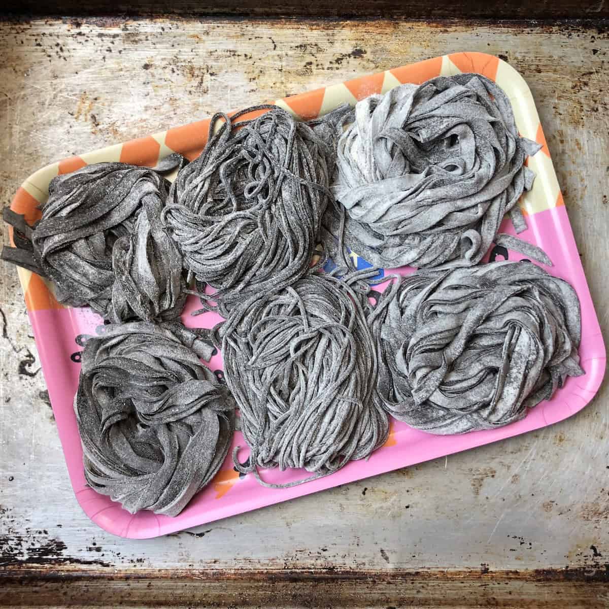 Six nests of fresh squid ink pasta including tagliatelle reginelle, black spaghetti, and fettuccine on semolina-dusted tray ready to freeze showing how to store homemade cuttlefish ink pasta.
