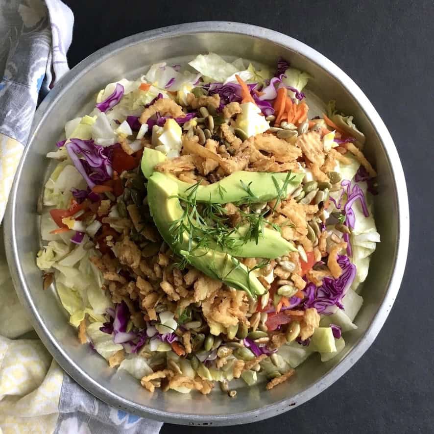 A loaded iceberg lettuce salad with sliced avocado, toasted sesame seeds, crispy french fried onions, shredded carrots, purple cabbage, cucumber, toasted pumpkin seeds, diced tomatoes, and chopped hard boiled eggs. A protein-packed meatless lenten salad.