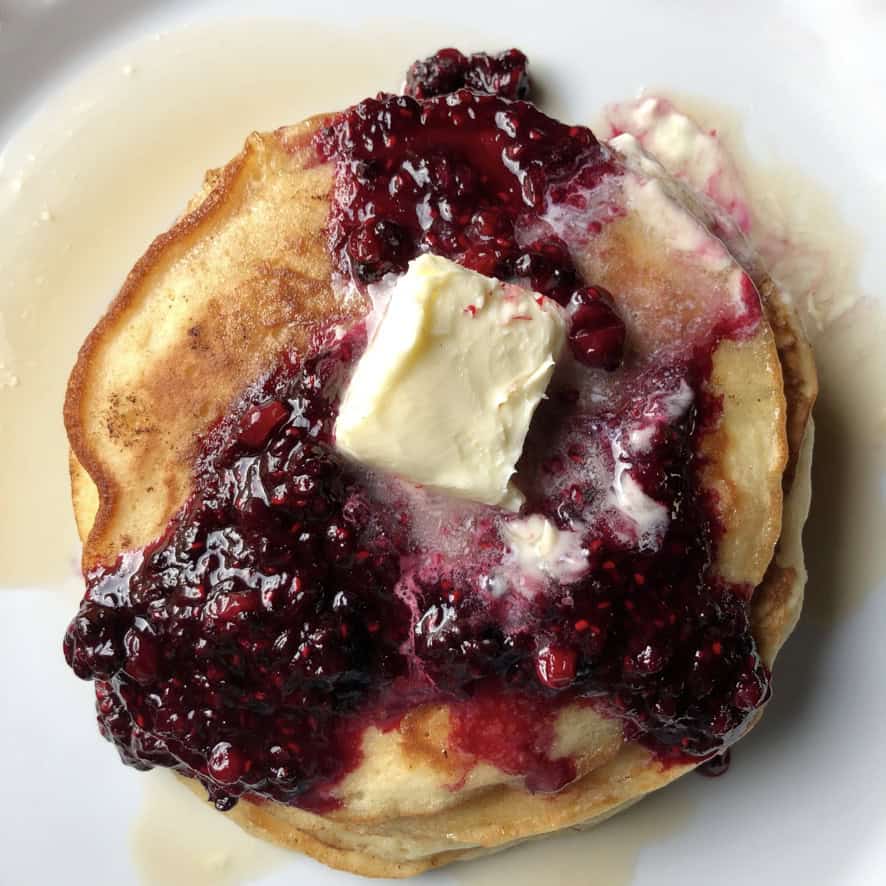 A tall stack of light and fluffy pancakes drizzled with maple syrup and homemade berry sauce with a pat of butter on top, ready to eat. A perfect meatless lenten breakfast for dinner idea.