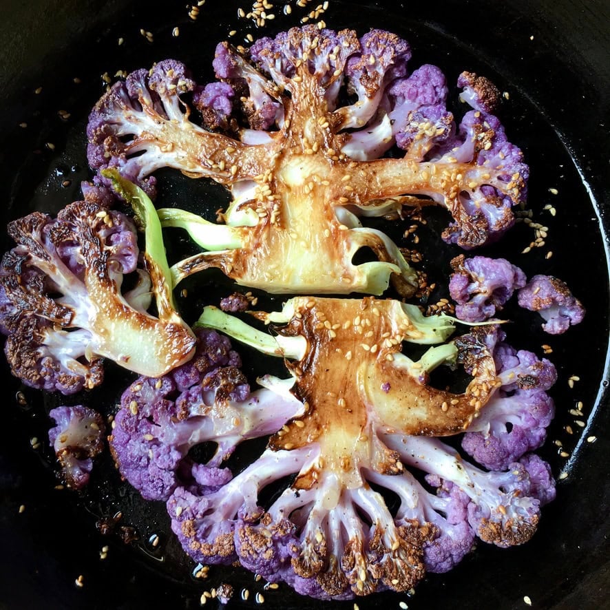 Pan-seared purple cauliflower steaks crusted with sesame seeds in a cast iron skillet, showing golden caramelized edges and vibrant purple color. A stunning meatless lenten side dish or vegetarian main.