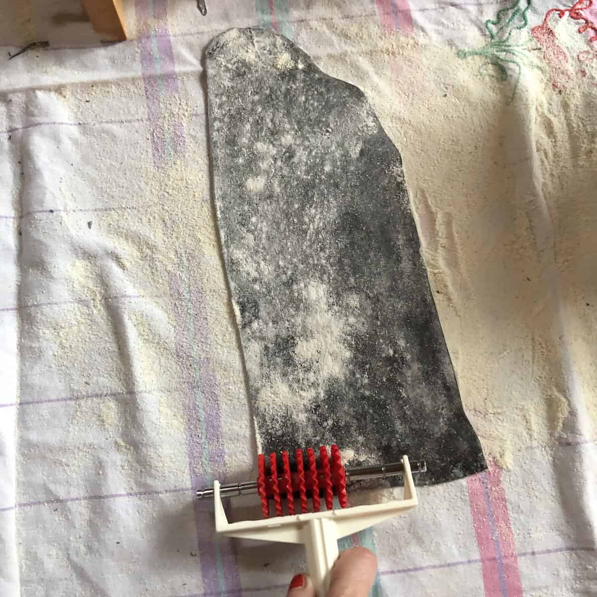 My hand using an Italian reginelle zigzag pasta cutter (aka Pasta bike) to make zigzag edge black tagliatelle from squid ink pasta sheet on floured surface.