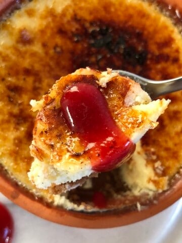 White chocolate crème brûlée cheesecake with Biscoff cookie crust and homemade strawberry coulis, showing a spoonful removed from the ceramic ramekin with a drizzle of strawberry sauce on top for Valentine's Day cheesecake recipes or romantic desserts for two.
