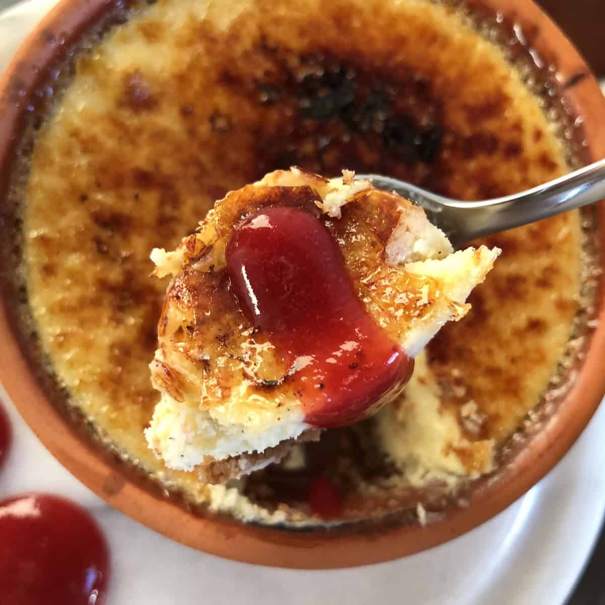 White chocolate crème brûlée cheesecake with Biscoff cookie crust and homemade strawberry coulis, showing a spoonful removed from the ceramic ramekin with a drizzle of strawberry sauce on top for Valentine's Day cheesecake recipes or romantic desserts for two.