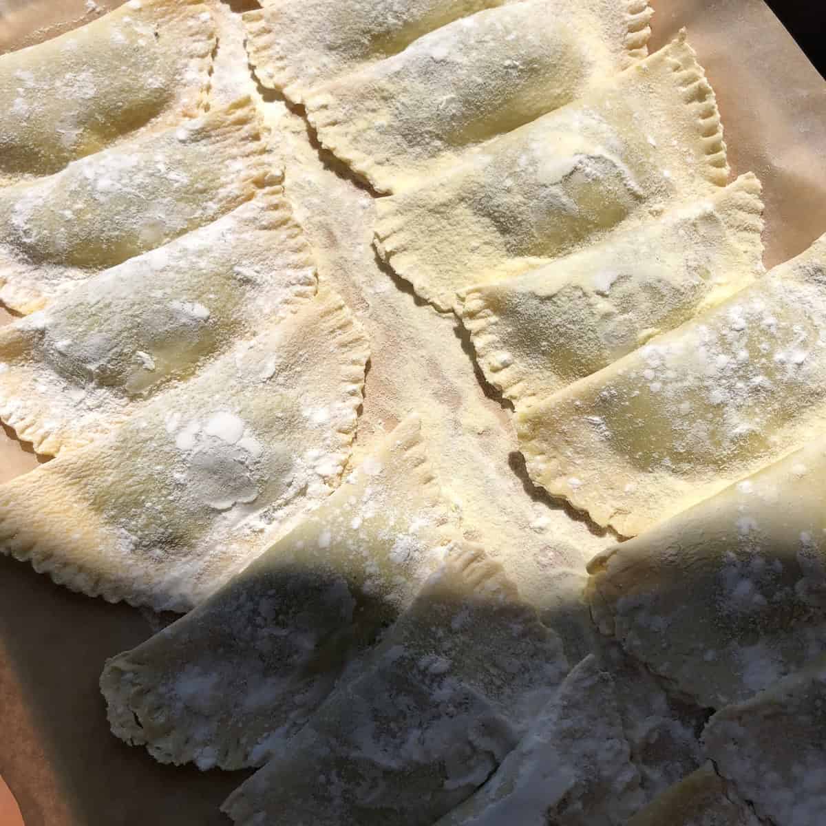 A tray of homemade Easter mezzaluna spinach and ricotta ravioli on semolina-dusted parchment, dusted with 00 flour and covered with a linen towel while resting - perfect for making ravioli ahead of time.