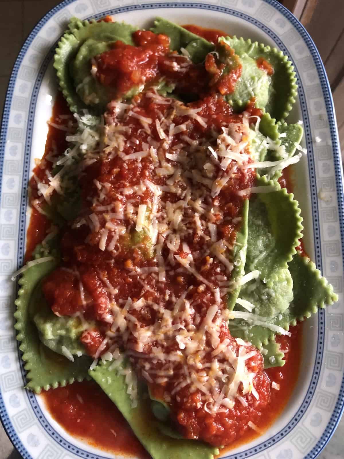 A large ceramic oval serving platter piled high with homemade green spinach and ricotta ravioli covered in homemade tomato sauce and freshly grated cheese — the ultimate vegetarian ravioli centerpiece.