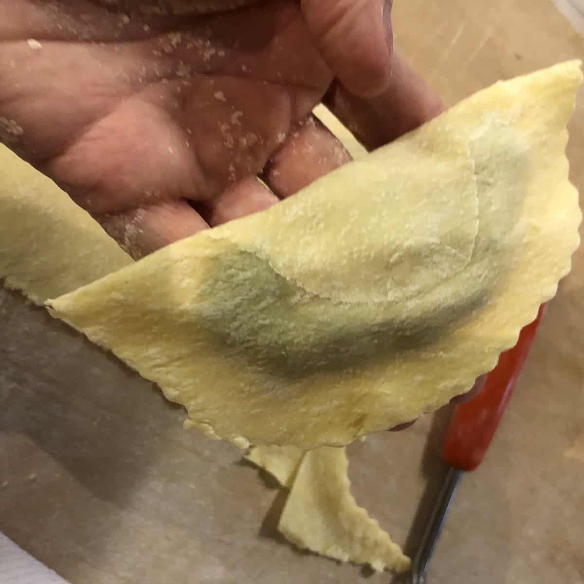 Nonna Anna holding up a just-cut mezzaluna spinach and ricotta ravioli before pressing and sealing, showing the traditional homemade spinach and ricotta ravioli method.