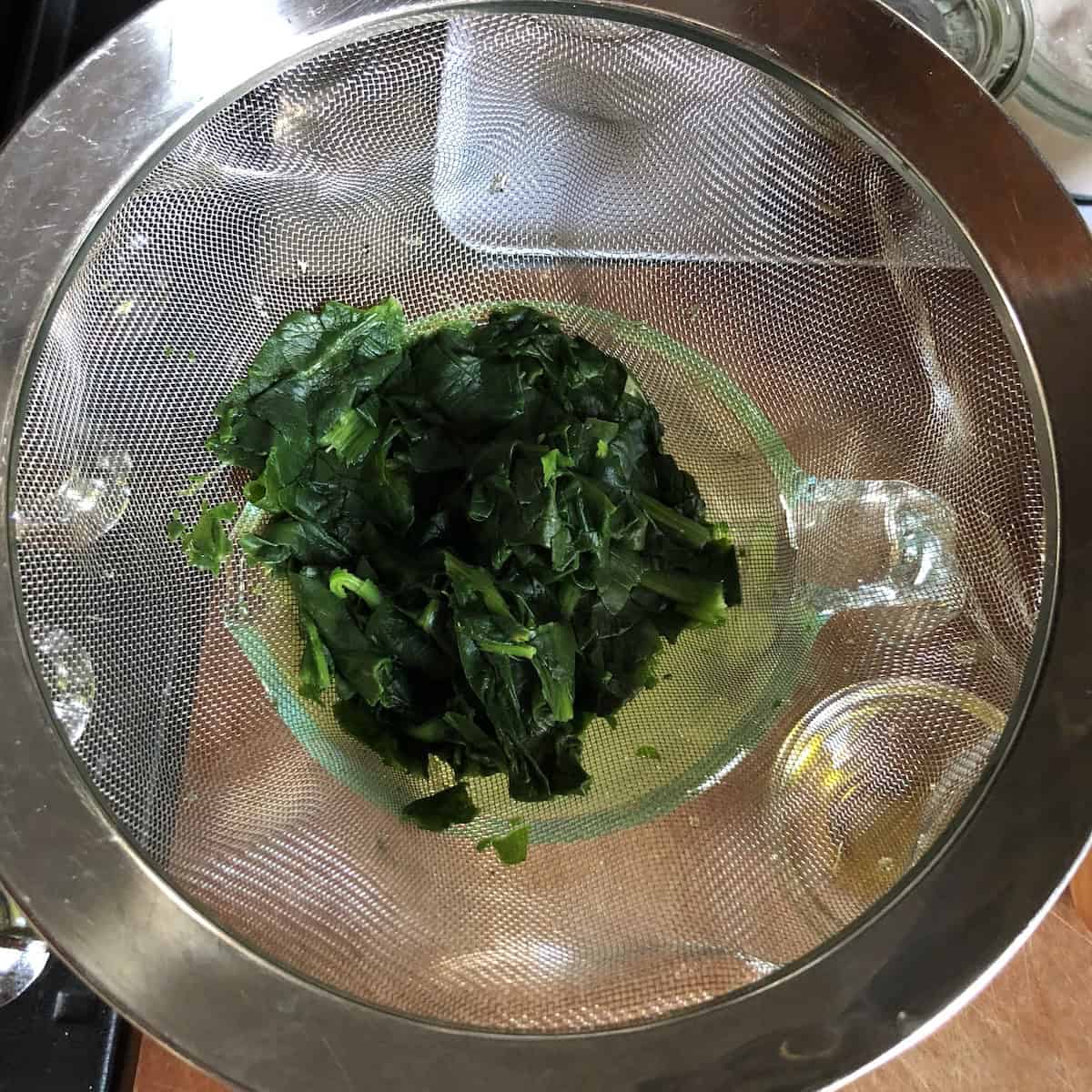 Blanched fresh spinach being strained in a fine mesh sieve to remove excess water when using fresh spinach for homemade fresh ravioli filling.
