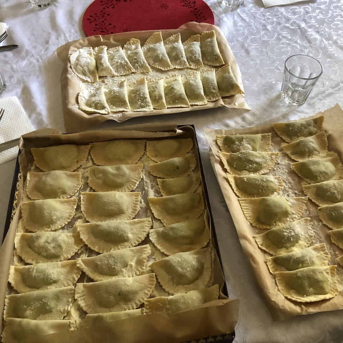 A table filled with three trays of homemade spinach and ricotta ravioli made by hand with Nonna Anna in her Northern Italian kitchen, proof that homemade ravioli can be made in advance for Easter.