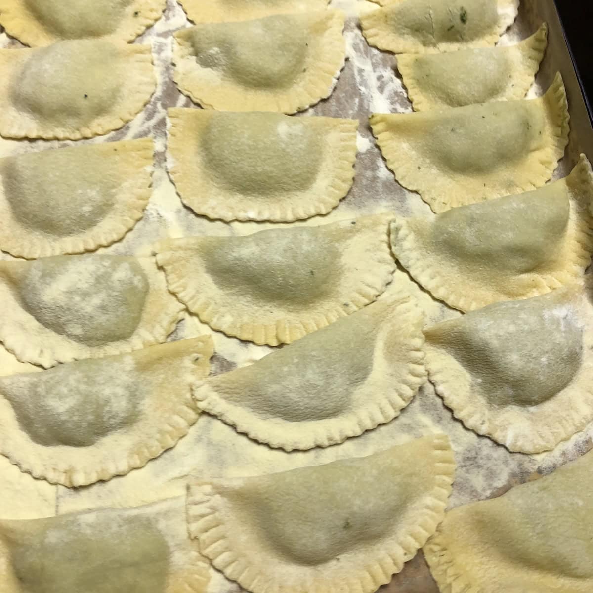 A tray filled with perfect Easter mezzaluna spinach and ricotta ravioli on a semolina-dusted parchment-lined sheet tray, ready to cook or freeze for a homemade spinach ravioli recipe.