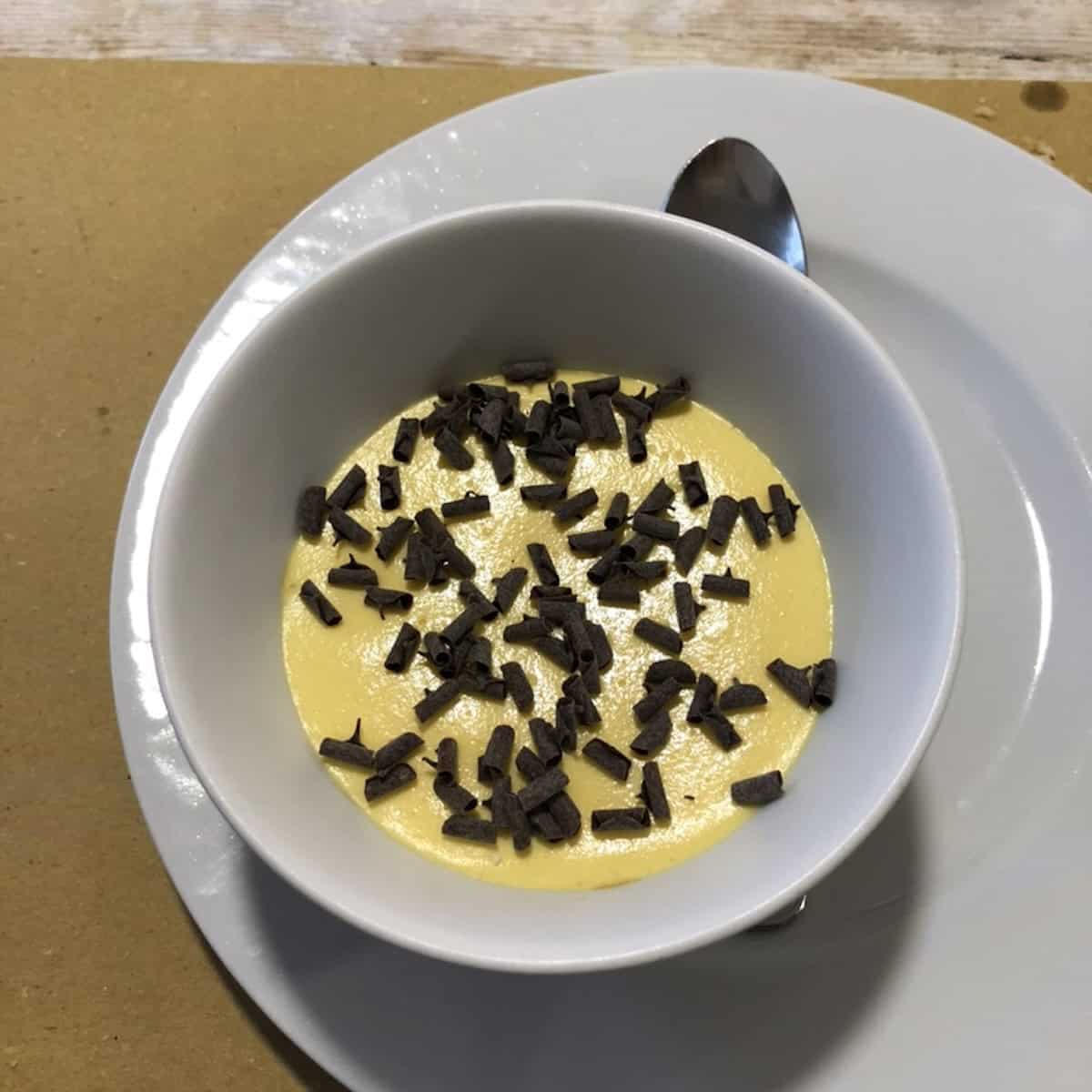 Individual osteria tiramisu served in a white ceramic cappuccino mug on a saucer with dark chocolate curls and no cocoa powder, showing an authentic Italian restaurant dessert.