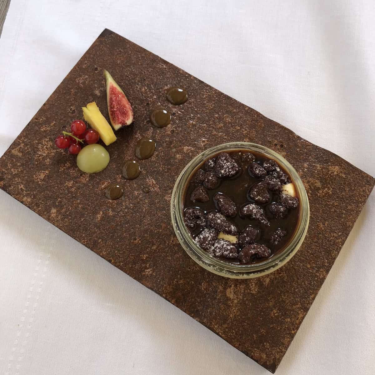 Fine dining Italian restaurant tiramisu in a glass jar with cocoa powder, chocolate gravel, and espresso poured on top, plated with chocolate sauce, red currants, and fresh fruit. 
