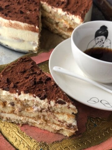 Slice of perfect tiramisu next to a Polspotten mug with handpainted woman holding her nose under the espresso, with the larger tiramisu in the background.