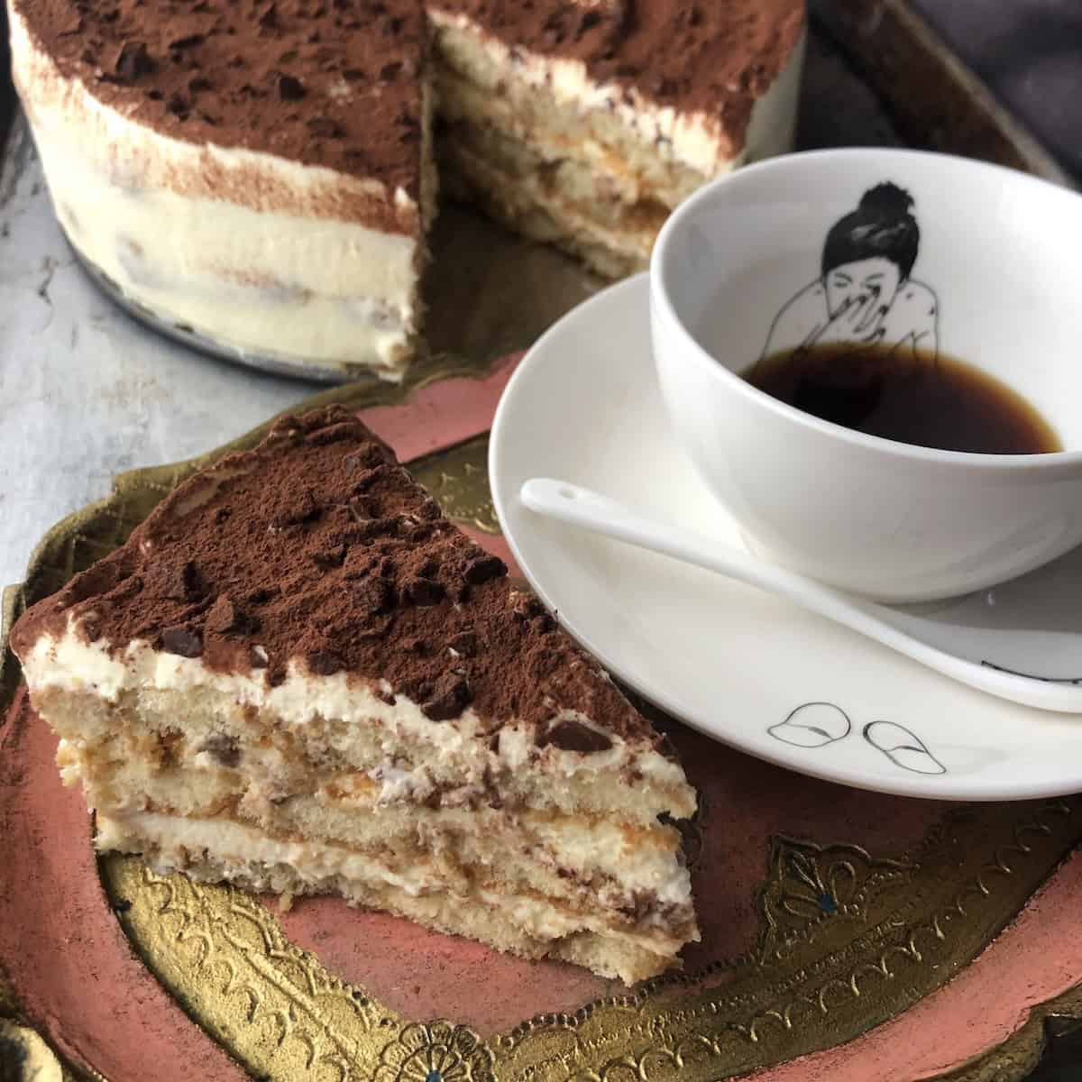Slice of perfect tiramisu next to a Polspotten mug with handpainted woman holding her nose under the espresso, with the larger tiramisu in the background.