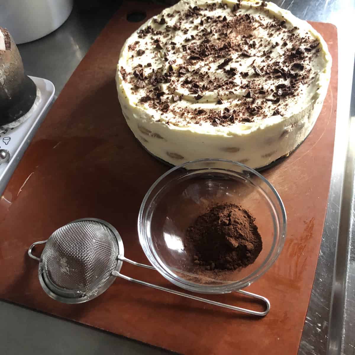 Cocoa powder next to a small sieve and chilled tiramisu topped with chopped 78 percent dark chocolate before dusting.
