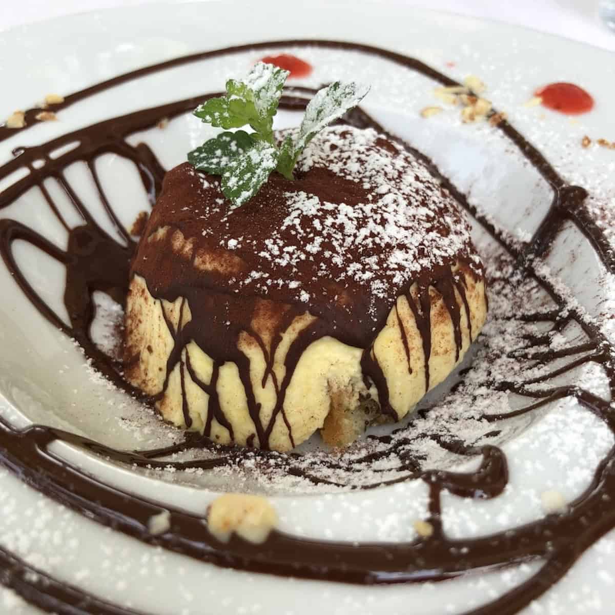Fine dining Italian restaurant tiramisu unmolded in a mound shape with hardened chocolate, cocoa powder, powdered sugar, mint sprig, chocolate sauce drizzle, strawberry coulis dots, and toasted hazelnuts.
