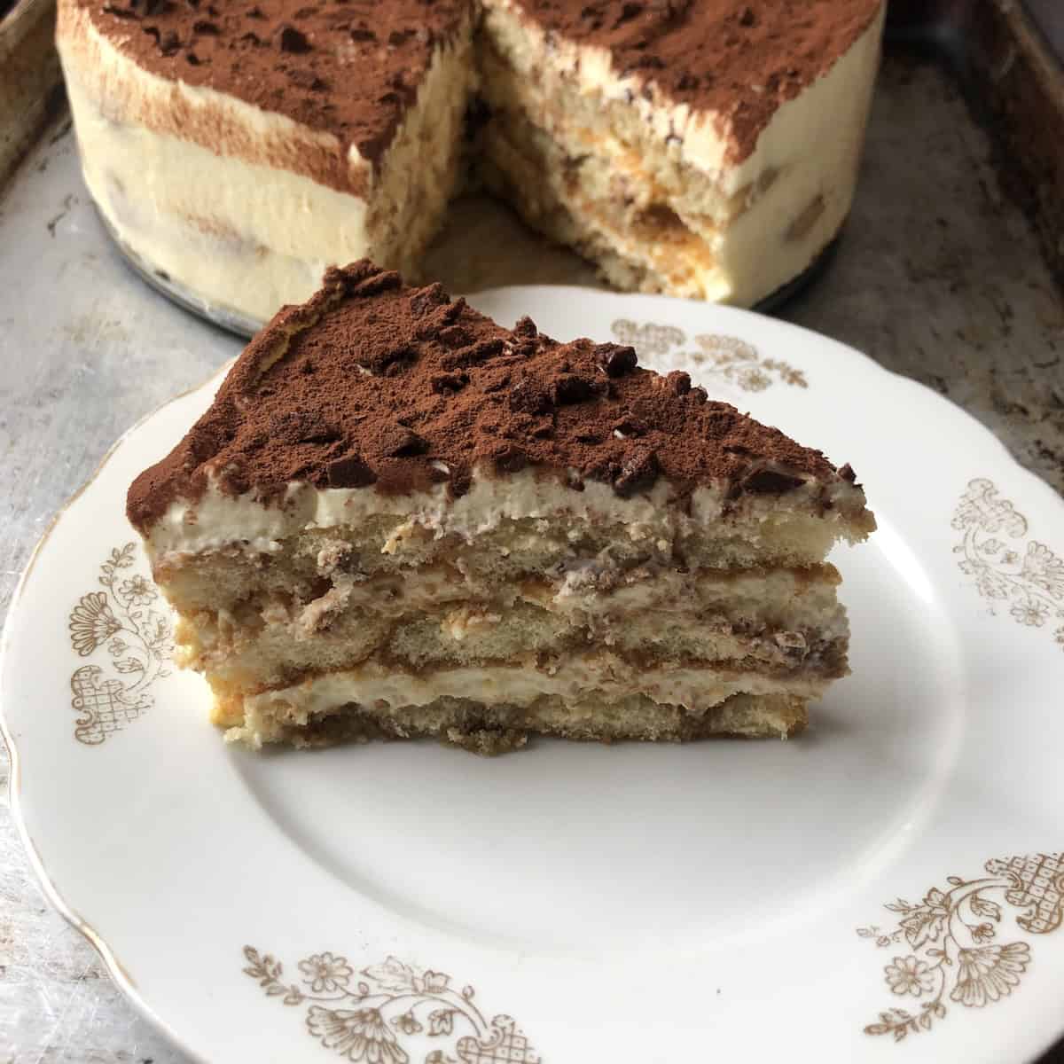 Slice of tiramisu on a dessert plate in front of the full tiramisu showing creamy mascarpone layers and perfectly espresso soaked ladyfingers dusted with cocoa powder and dark chocolate, Italian tiramisu recipe.