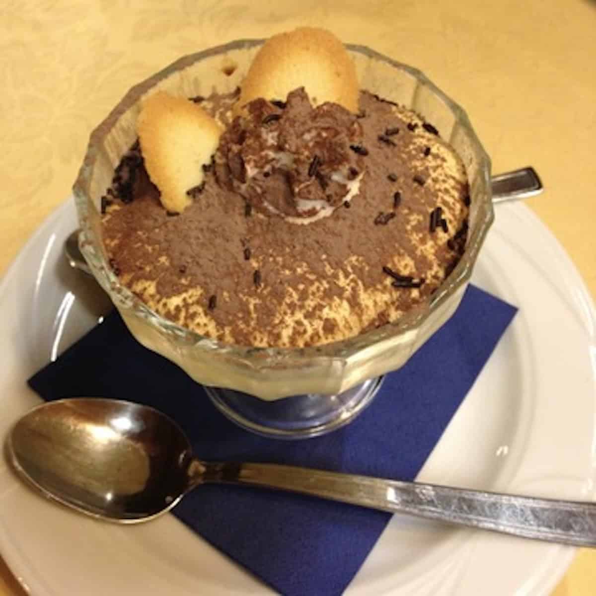 Trattoria tiramisu served in a dessert coupe set atop a blue napkin resting on a plate with two spoons, cocoa powder, chocolate sprinkles, and tuile cookies as garnish.
