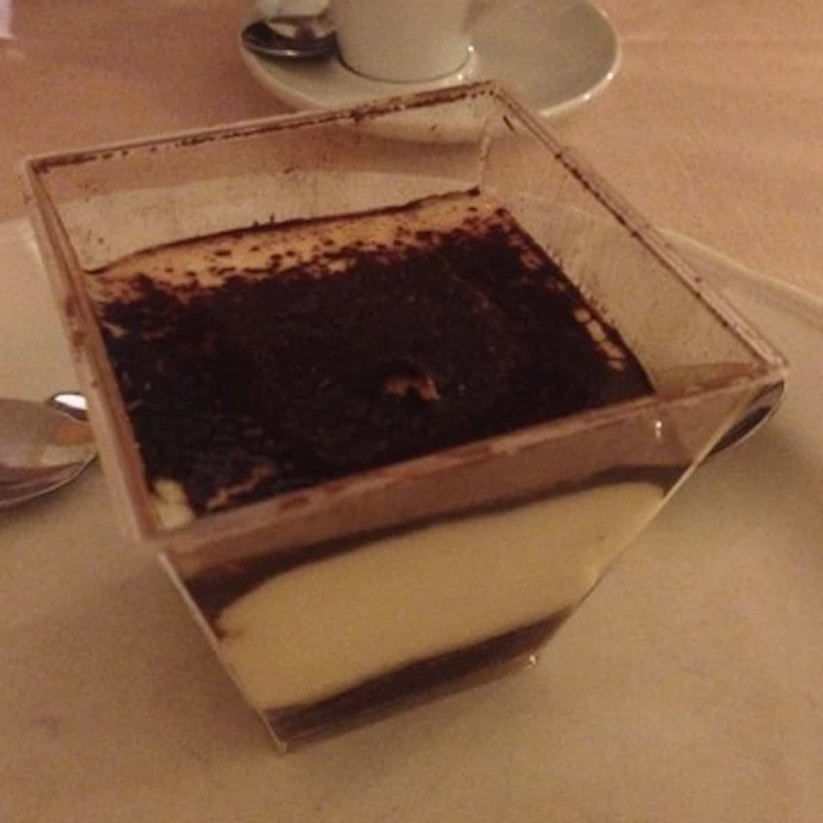 Italian restaurant tiramisu in a square individual serving with espresso at the bottom, alternating layers of mascarpone cream and ladyfingers topped with cocoa powder.
