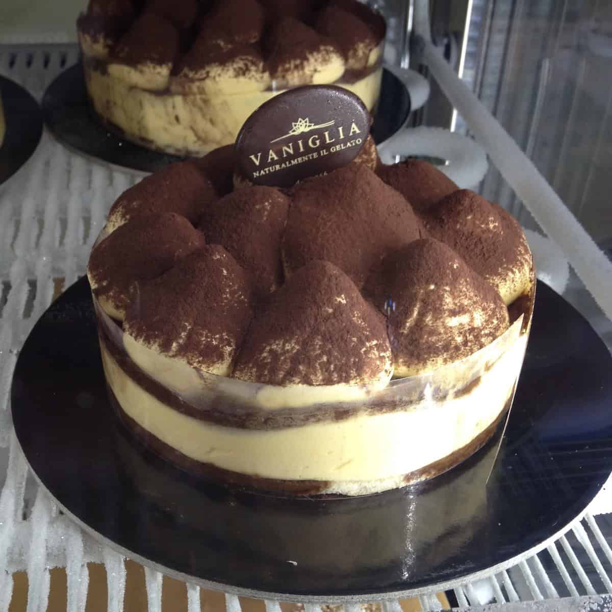 Whole tiramisu from a local Italian pasticceria with acetate-lined cake shape, drop shaped mascarpone cream on top dusted with cocoa powder and shaved chocolate curls.